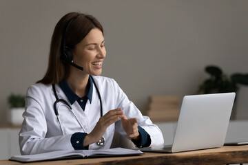 Happy young female doctor therapist general practitioner in headset with mic looking at computer screen, holding distant video call meeting giving consultation to client, telemedicine concept.