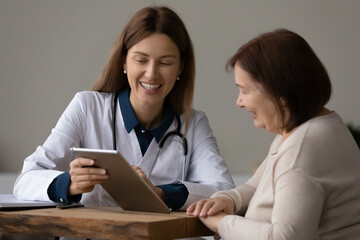 Happy young female GP doctor physician showing health test results on digital computer tablet to happy old mature retired woman, feeling satisfied with disease treatment, medicine and modern tech.