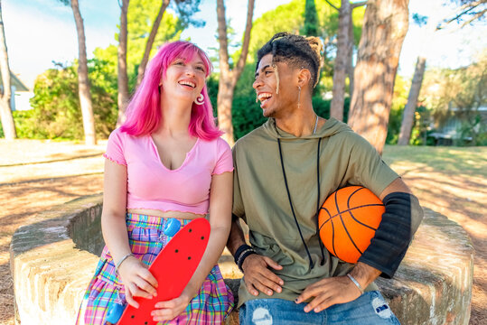 Alternative Diverse Couple Hanging Out Together Chilling And Smiling Sitting In A Park Holding A Basketball And Skateboard. Happy Interracial Friends Laughing Outdoors. Joy, Fun And Lifestyle Concept