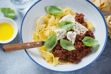 Closeup of italian pappardelle pasta served with bolognese sauce, mozzarella and fresh green basil in a white plate, selective focus