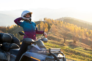 Happy female driver in protective helmet enjoying offroad riding on ATV quad motorbike in autumn mountains at sunset.