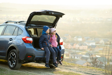 Side view of sister and brother sitting in car trunk and looking on nature. Concept of resting on fresh air with family.