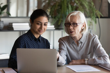 Serious mature businesswoman mentor coach in glasses teaching Indian woman intern student, using...