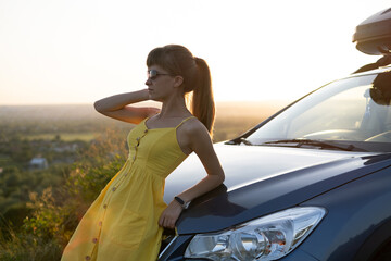 Happy young woman in yellow dress standing near her vehicle looking at sunset view of summer nature. Travelling and vacation concept.