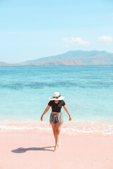 Woman tourist in summer hat walking on pink sandy beach with sea view and hills at Labuan Bajo