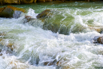 Mountain river with small waterfall with clear turquoise water falling down between wet boulders with thick white foam.