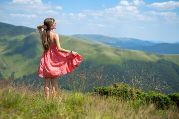 Young woman in red dress standing on grassy field on a windy day in summer mountains enjoying view of nature.