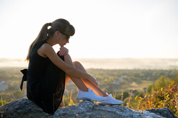 Young depressed woman in black short summer dress sitting on a rock thinking outdoors at sunset....