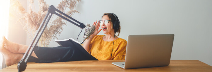 Woman radio host recording and broadcasting her podcast from homemade studio. Female podcaster talk into microphone on table. Wide image.