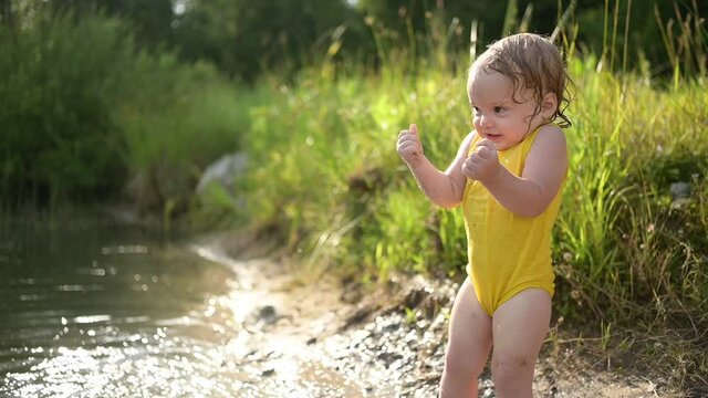 Little funny cute blonde girl child toddler in yellow wet bodysuit playing by the lake waterside shore at sunset outside. Baby with mud on hands and legs. Water activities at summer