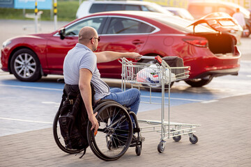 Fototapeta na wymiar Adult disabled man in a wheelchair pushes a cart towards a car in a supermarket parking lot
