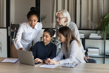 Diverse colleagues with mature businesswoman mentor wearing glasses discussing online project at...