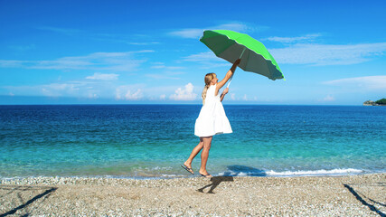 Young girl in a white dress with a green umbrella in her hands is raised by the wind over a pebble...