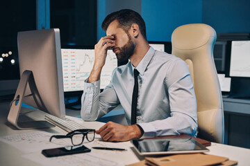 Frustrated young man in shirt and tie massaging nose and keeping eyes closed while staying late in the office