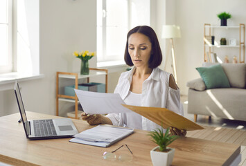 Serious woman sitting at desk in her home office, looking through business documents, doing...