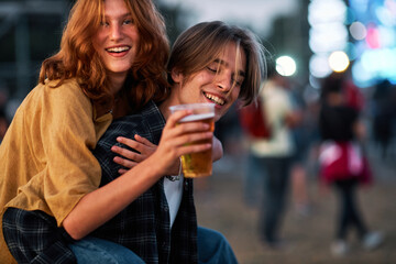 A young couple is having a piggyback ride at a rock music festival. Hippie girl holding a plastic glass of beer and having a good time.