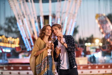 A young, happy couple is standing at a fair outdoors, chatting and drinking beer.