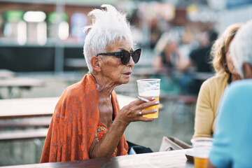 An old woman with sunglasses is sitting with her friends at a music festival, chatting, drinking...