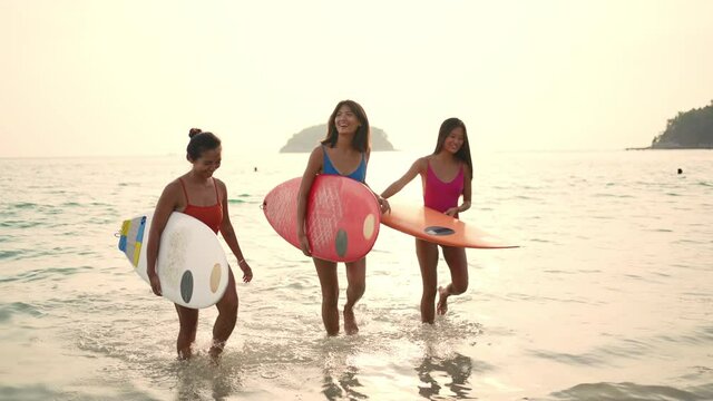 Confidence Asian woman girl friends in swimwear holding surfboard and walking together on the beach at summer sunset. Female friendship enjoy outdoor activity lifestyle play extreme sports surfing