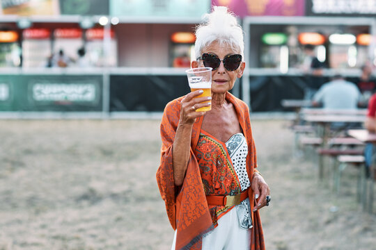 A grandma standing outdoor at a music festival and showing her beer. She is having a good time.