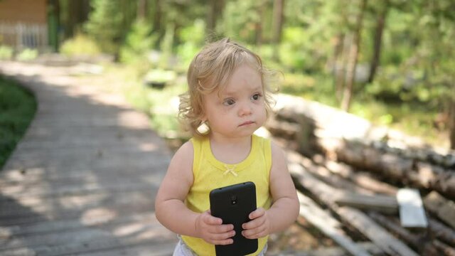 Little funny cute blonde girl child toddler holding playing big black smartphone outside at summer at countryside. Healthy happy childhood concept. Kids and technologies