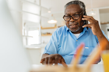An old black man is sitting at his home office, talking on the phone with a customer support worker and using his laptop. - 453604297