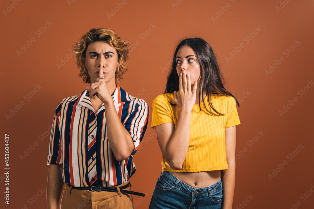 Wall mural Stylish caucasian man with a hushing gesture and a young surprised female against a brown background