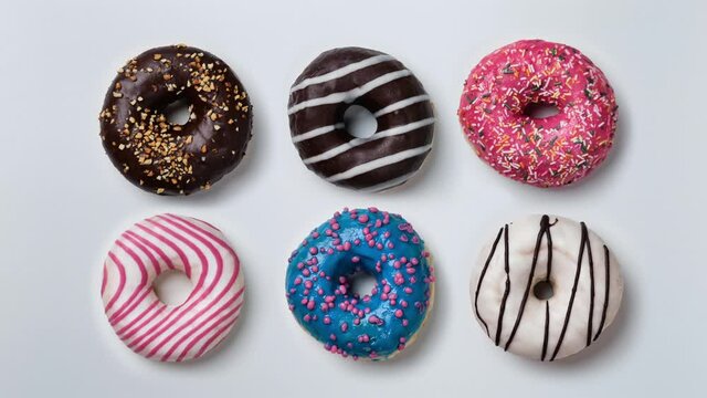 Donuts of different colors changing at white background. Stop motion. Six glazed sweet donuts. Bakery and food concept. Top view. Various colorful donuts. Chocolate, pink, blue donuts in 4K, UHD