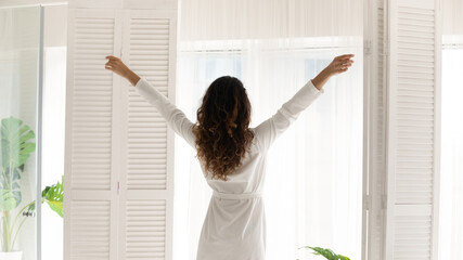 Fototapeta na wymiar Back rear view young slim woman with brown curly wavy long hair in robe standing near window, stretching muscles, welcoming new day, enjoying carefree vacation weekend good morning time at home.