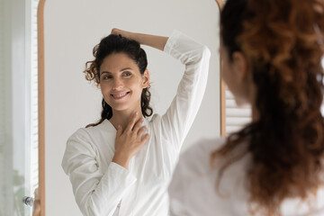 Smiling attractive millennial hispanic latina woman in bathrobe looking in big mirror, feeling...