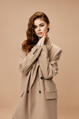 young brunette in coat holds hands near face on beige background cropped view