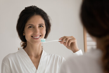 Mirror reflection happy young beautiful woman in bathrobe holding toothbrush with applied paste, looking at camera, starting morning hygienic oral healthcare dental procedure, cleaning teeth.