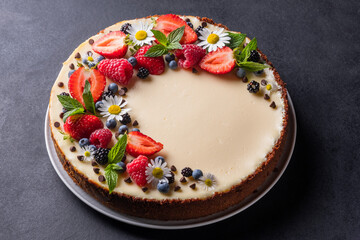 Cheesecake pie with fresh berries, strawberries, blueberries and blackberries and mint chamomiles in a round plate on a black background.