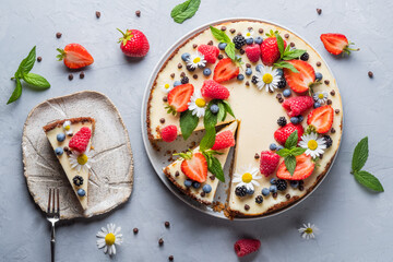 Cheesecake pie with fresh berries, strawberries, blueberries and blackberries and mint chamomiles in a round plate on a grey background.