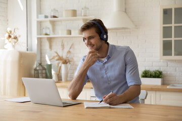 Smiling millennial generation male student in wireless headphones listening educational lecture online on computer, writing notes in copybook improving knowledge distantly, enjoying e-learning.