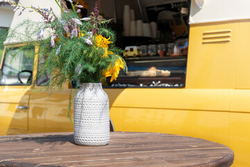 Yellow street coffee shop on wheels near table with flowers. Vintage food truck with empty round table on a sunny day.
