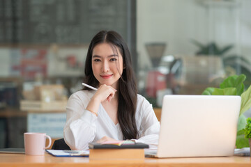 Fototapeta premium Happy young Asian girl working at a coffee shop with a laptop.Woman happy smiling to camera in cafe day.
