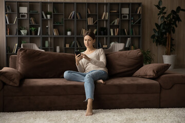 Full length woman holding smartphone, sitting on couch at home, attractive young female looking at phone screen, chatting in social networks with friends, shopping, browsing mobile device apps