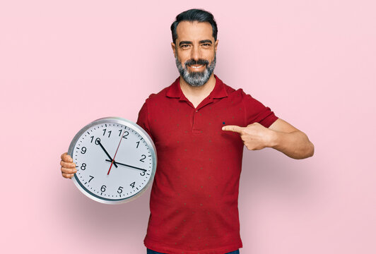 Middle aged man with beard holding big clock pointing finger to one self smiling happy and proud