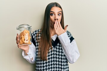 Beautiful brunette young woman holding jar with spaghetti pasta covering mouth with hand, shocked...