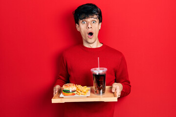 Handsome hipster young man eating a tasty classic burger with fries and soda afraid and shocked...