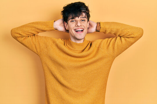 Handsome hipster young man wearing casual yellow sweater relaxing and stretching, arms and hands behind head and neck smiling happy