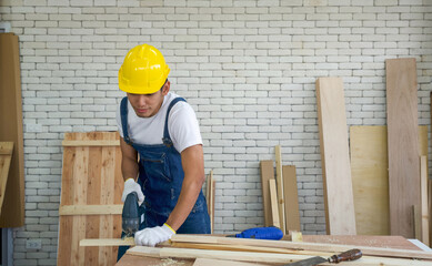 Asian carpenter wearing a yellow hardhat, use electric hand saw to cut plywood at the woodworking facility. A desk full of hand tools and wood piles. Morning work atmosphere in the workshop room.