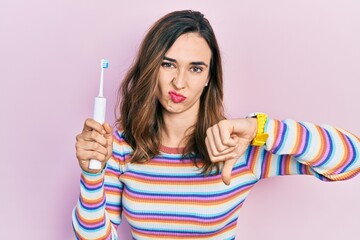 Young hispanic girl holding electric toothbrush with angry face, negative sign showing dislike with thumbs down, rejection concept