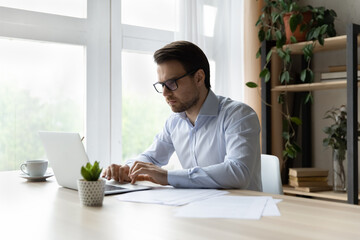 Serious millennial businessman working at laptop in office. Focused young man in glasses, employee, lawyer using computer at workplace, typing, chatting online, watching virtual presentation