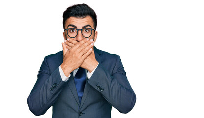 Hispanic man with beard wearing business suit and tie shocked covering mouth with hands for...