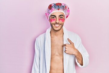 Young hispanic man wearing bathrobe and shower cap using eyes bags patches pointing finger to one self smiling happy and proud