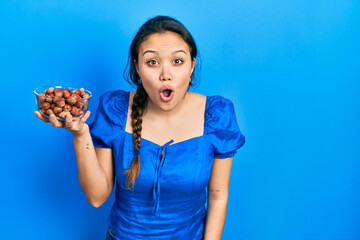Young hispanic girl holding bowl of chestnuts scared and amazed with open mouth for surprise, disbelief face