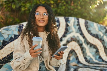 Young latin woman using smartphone and drinking coffee sitting on the bench.