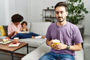 Hispanic father of interracial family drinking a cup coffee relaxed with serious expression on face. simple and natural looking at the camera.